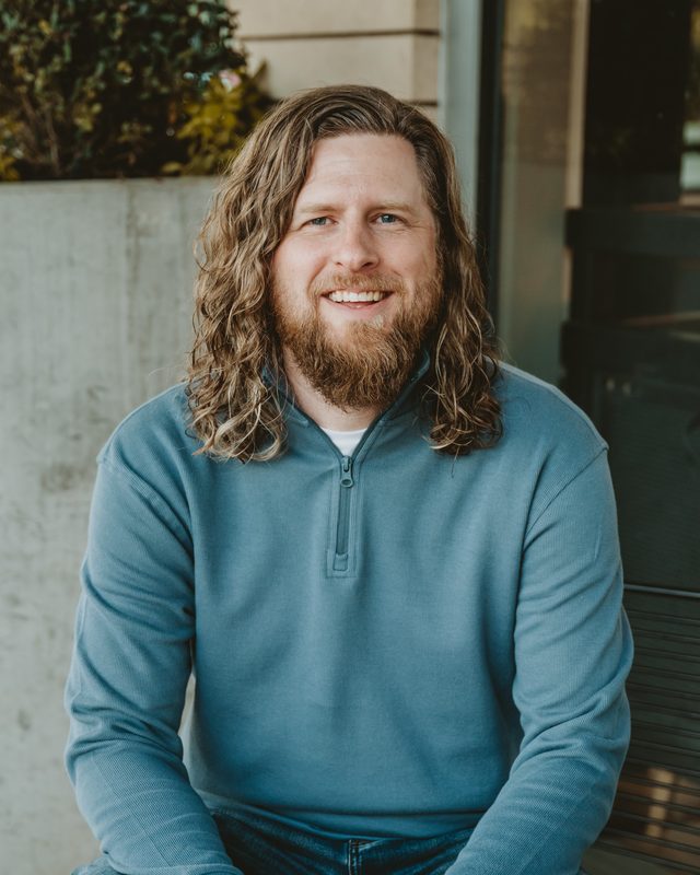 Daniel Heath Ferguson, MA, LPC, smiling, seated outdoors in a blue quarter-zip